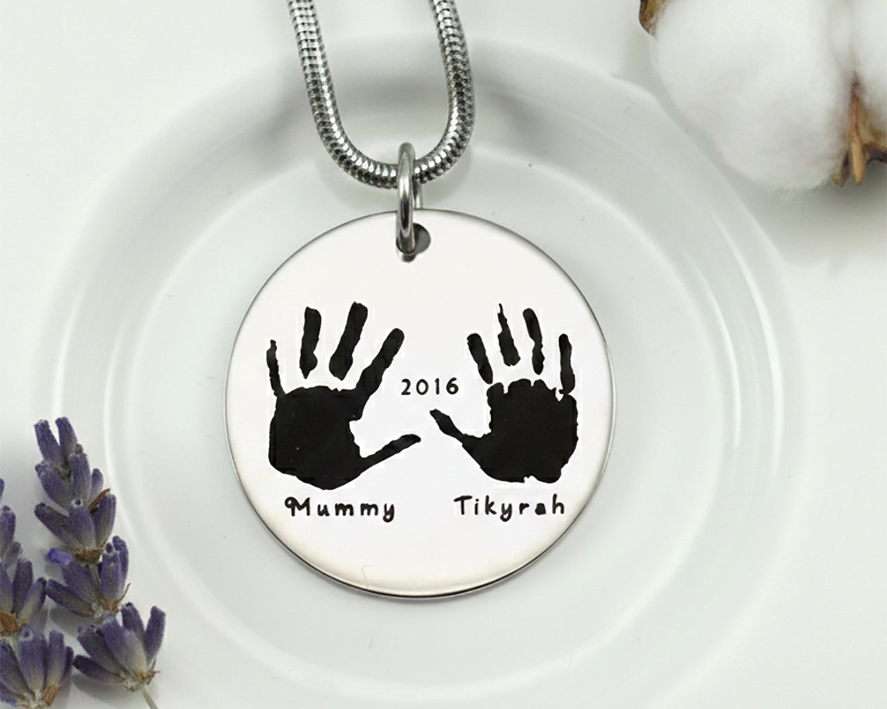 Belle Fever Hand on My Heart Necklace — silver round pendant engraved with handprints, displayed on a soft surface, a meaningful keepsake symbolising love and remembrance.